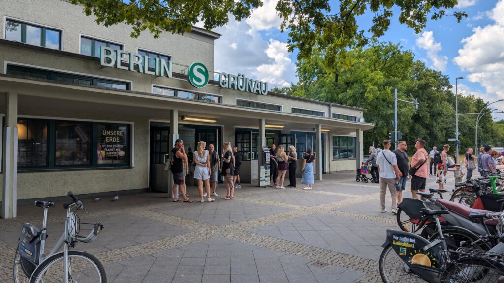 Eingang zum S-Bahnhof Grünau mit Fahrrädern und wartenden Menschen.